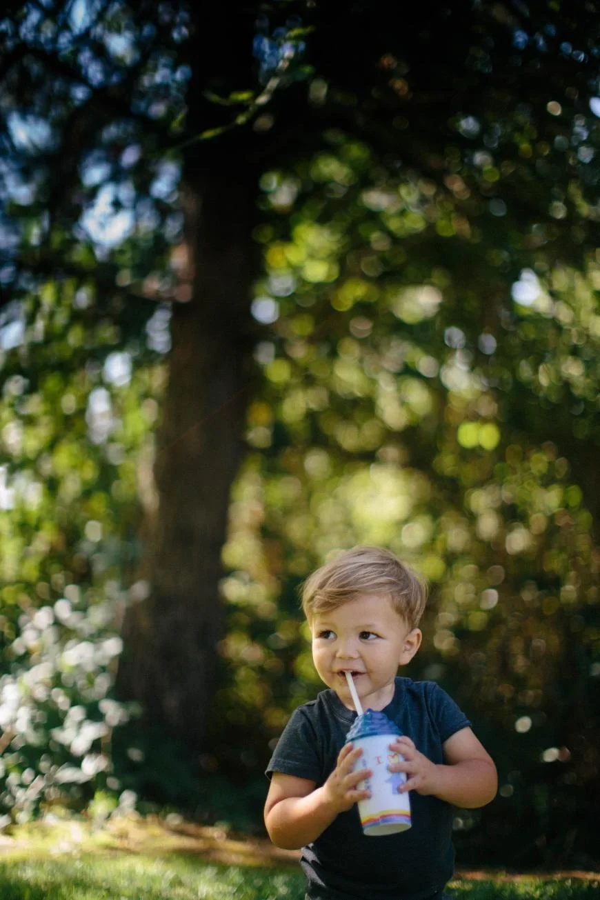 Young child drinking from a colorful cup with straw in a sunlit forest setting with bokeh effect in background.