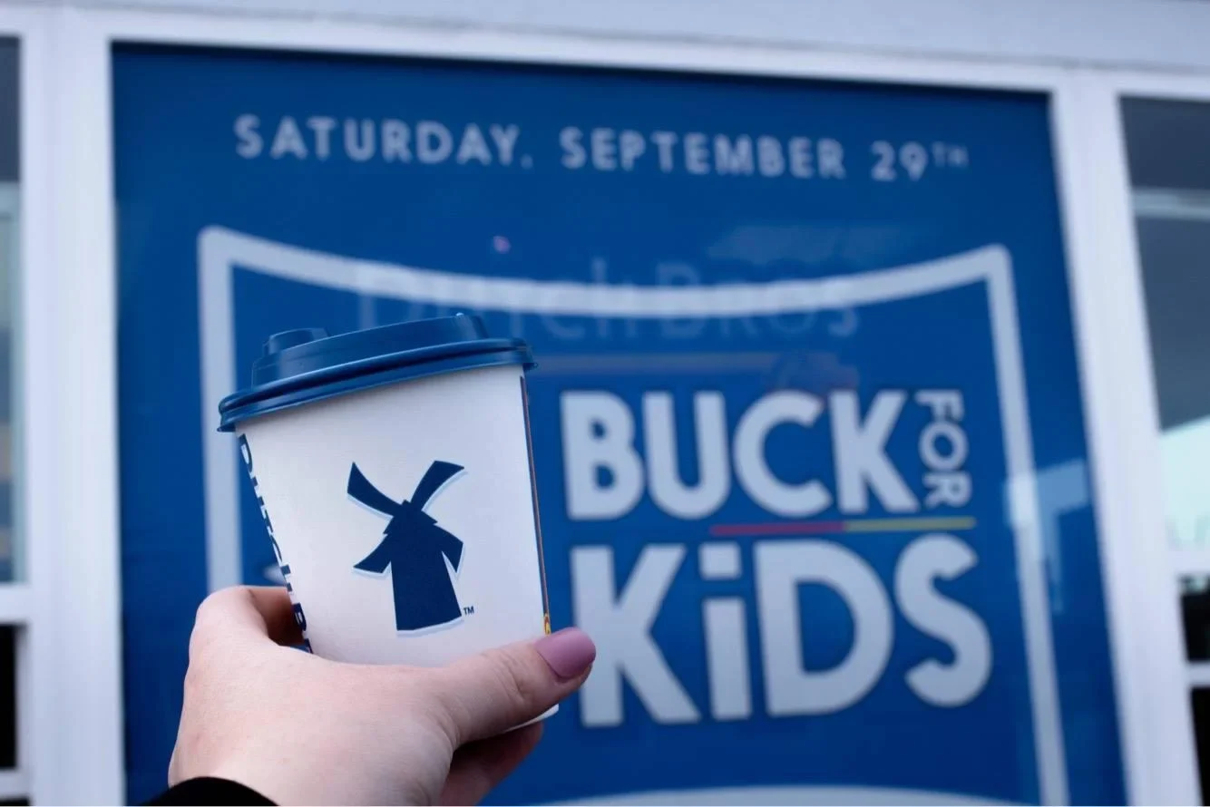 Hand holding Dutch Bros coffee cup in front of blue "Buck for Kids" event sign for Saturday, September 29th.