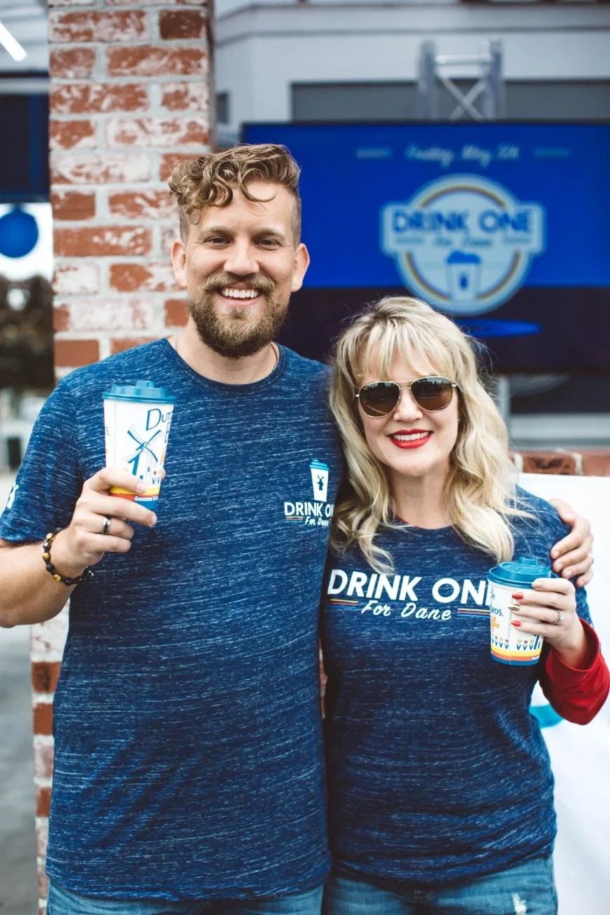 Two people in matching blue "DRINK ONE" shirts holding coffee cups outside a café with brick wall background.