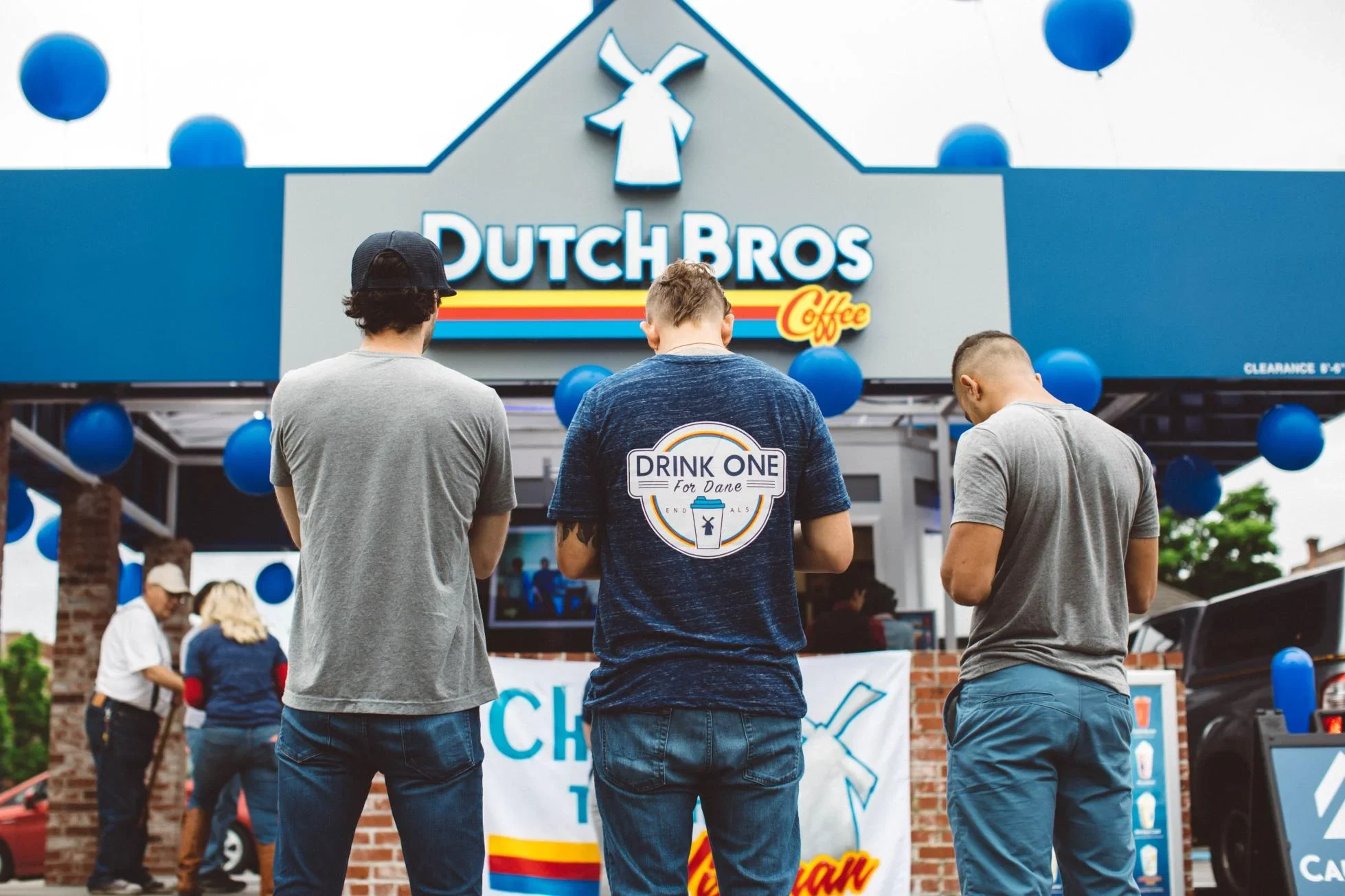 Three people facing Dutch Bros Coffee storefront decorated with blue balloons, one wearing a "Drink One for Dane" t-shirt.