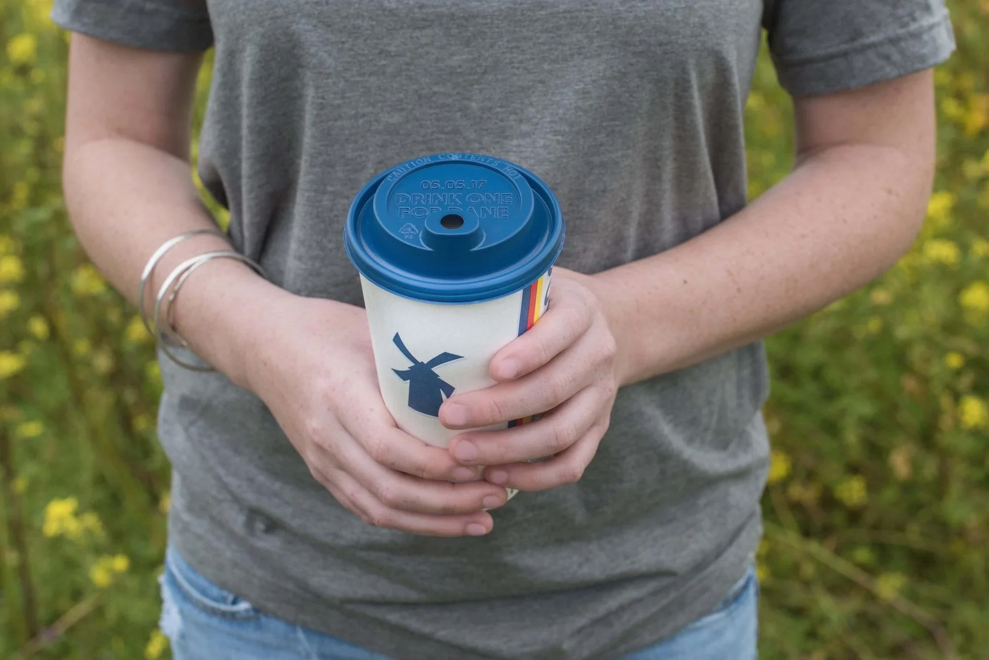 Person in gray shirt holding coffee cup with blue lid and windmill design against yellow flower background.